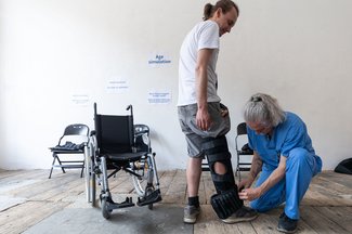 Links ein leerer Rollstuhl, hinten an der Wand ein Klappsessel. In der Mitte steht eine Person, der eben von einem rechts neben ihr knieenden Menschen in blauer Arbeitskleidung eine Beinschiene angelegt wird.