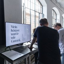 Rückenansicht von zwei Personen, die eine Videoinstallation und den Begleittext lesen.